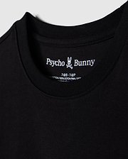 Close-up of a black t-shirt, showcasing the brand name "Psycho Bunny" and a small bunny skull logo inside the collar. Details include shirt size and material info, made from 100% Peruvian Pima cotton. The soft and smooth fabric highlights its premium quality. This particular piece is identified as the "KIDS LOS ANGELES CITY TEE - B0U861A2PC".