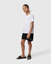 A young person stands casually wearing a white t-shirt, Psycho Bunny KIDS MALTA HYDROCHROMIC SWIM TRUNK - B0W321B2SW, and black sandals, facing slightly to their left, with a neutral expression and hands by their sides.