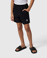 A person stands displaying Psycho Bunny hydrochromic swim trunks and black slide sandals, the trunks feature a small white logo on the left thigh. Only the lower half of the person is visible against a plain background.