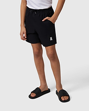 A person stands displaying Psycho Bunny hydrochromic swim trunks and black slide sandals, the trunks feature a small white logo on the left thigh. Only the lower half of the person is visible against a plain background.