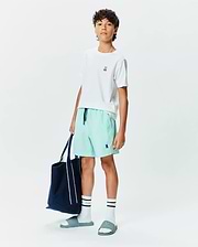 White classic fit jersey t-shirt with small embroidered Psycho Bunny logo, Kids Samos Hydrochromic Printed Swim Trunk in mint green with navy drawstring and embroidered logo, white crew socks with black stripes and Psycho text, and teal slides with embossed logo.
