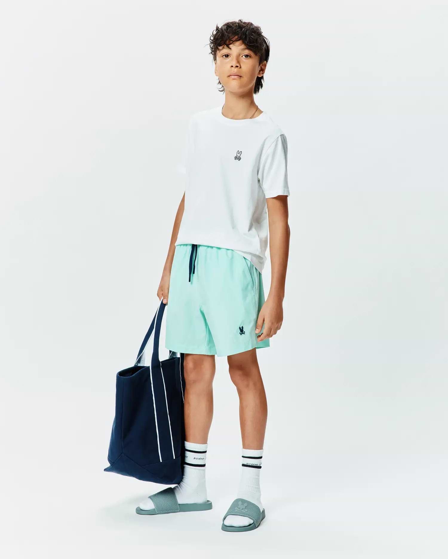 White classic fit jersey t-shirt with small embroidered Psycho Bunny logo, Kids Samos Hydrochromic Printed Swim Trunk in mint green with navy drawstring and embroidered logo, white crew socks with black stripes and Psycho text, and teal slides with embossed logo.