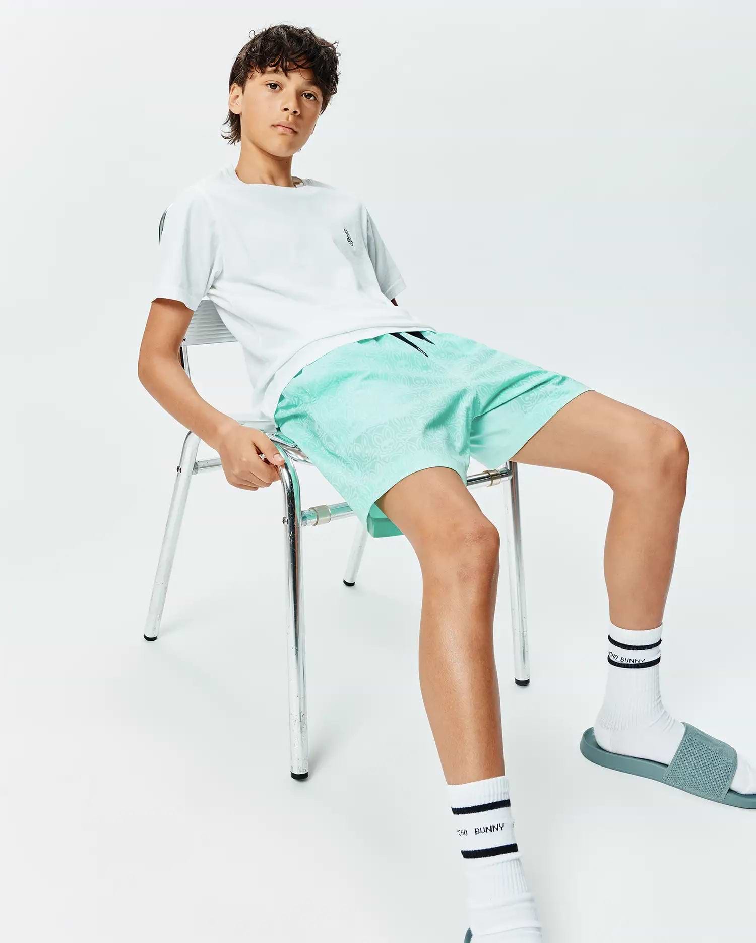 White t-shirt with small embroidered Psycho Bunny skull logo on chest, paired with Kids Samos Hydrochromic Printed Swim Trunk in mint green featuring a subtle all-over bunny skull pattern, and white Psycho Bunny socks.