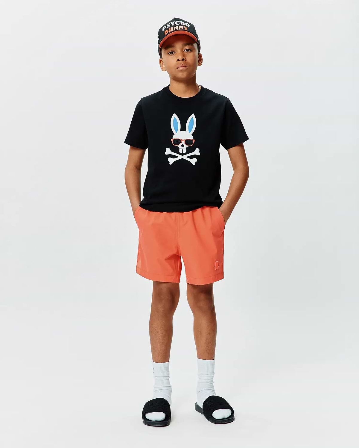 Black classic fit jersey t-shirt with large chest Psycho Bunny skull graphic, orange Kids Samos Hydrochromic Printed Swim Trunk with embroidered logo, and black Psycho Bunny baseball cap.