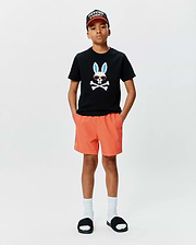 Black classic fit jersey t-shirt with large chest Psycho Bunny skull graphic, orange Kids Samos Hydrochromic Printed Swim Trunk with embroidered logo, and black Psycho Bunny baseball cap.
