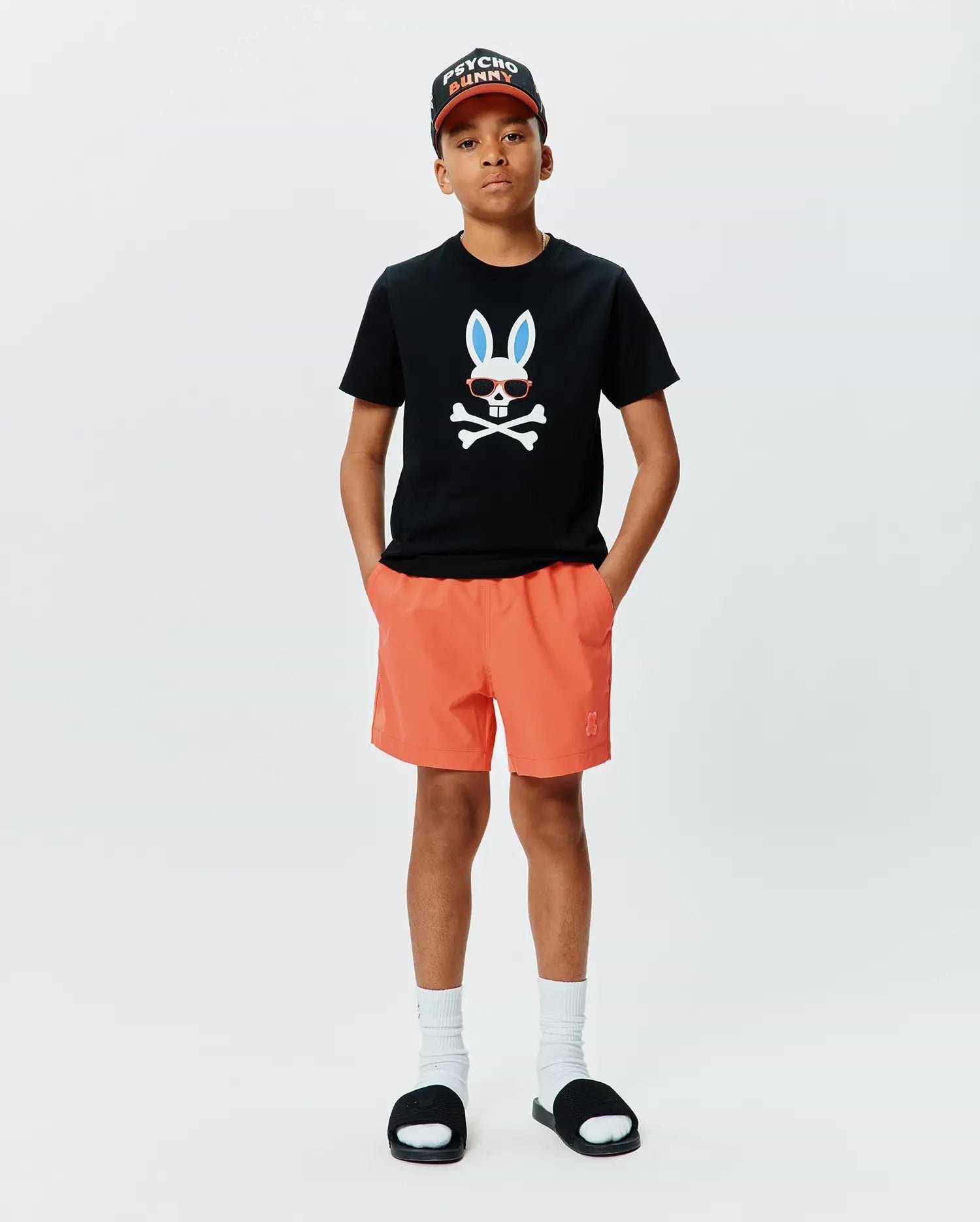 Black classic fit jersey t-shirt with large chest Psycho Bunny skull graphic, orange Kids Samos Hydrochromic Printed Swim Trunk with embroidered logo, and black Psycho Bunny baseball cap.