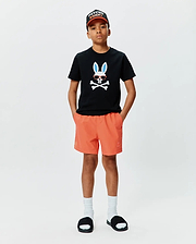 Black classic fit jersey t-shirt with large chest Psycho Bunny skull graphic, orange Kids Samos Hydrochromic Printed Swim Trunk with embroidered logo, and black Psycho Bunny baseball cap.