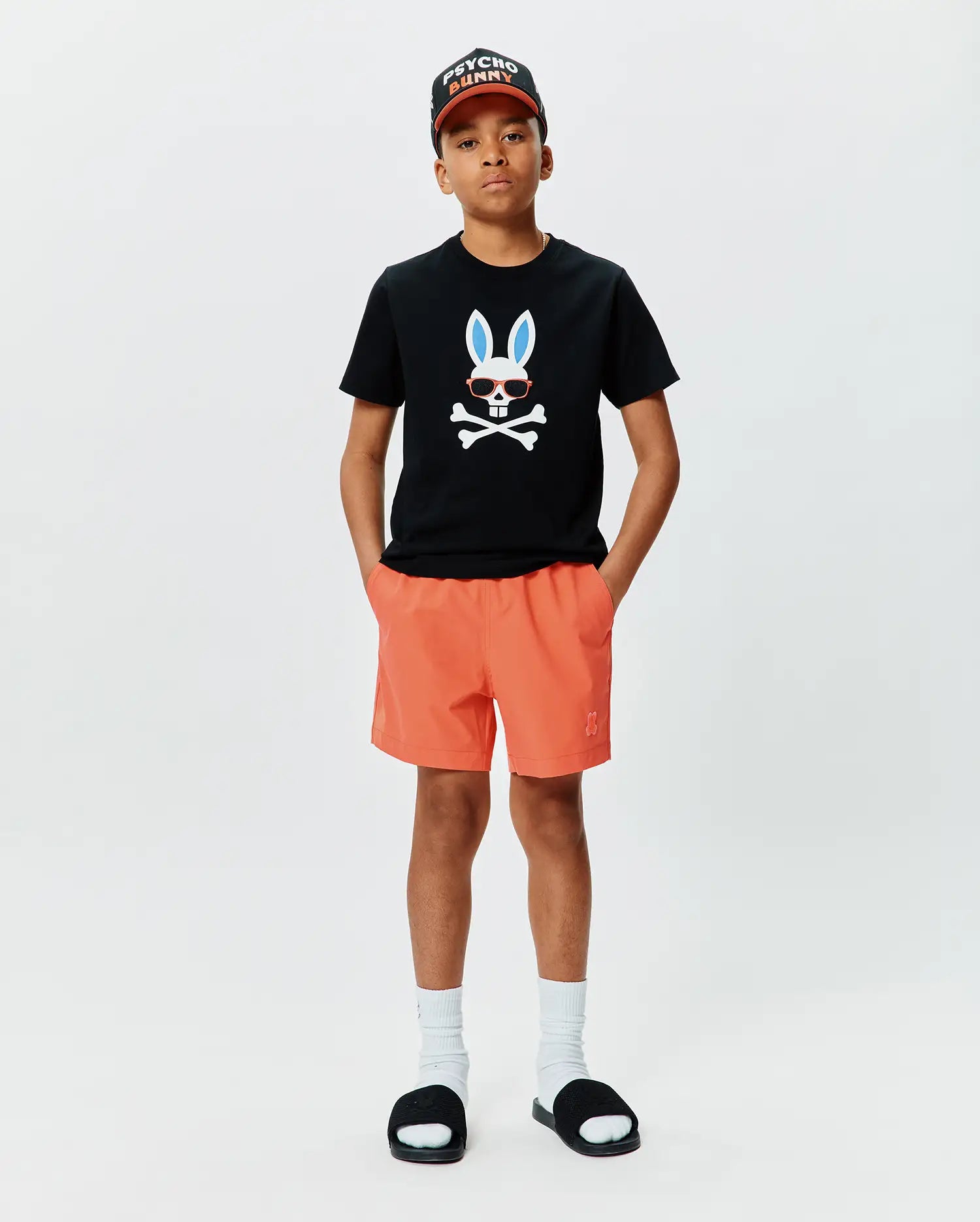 Black classic fit jersey t-shirt with large chest Psycho Bunny skull graphic, orange Kids Samos Hydrochromic Printed Swim Trunk with embroidered logo, and black Psycho Bunny baseball cap.