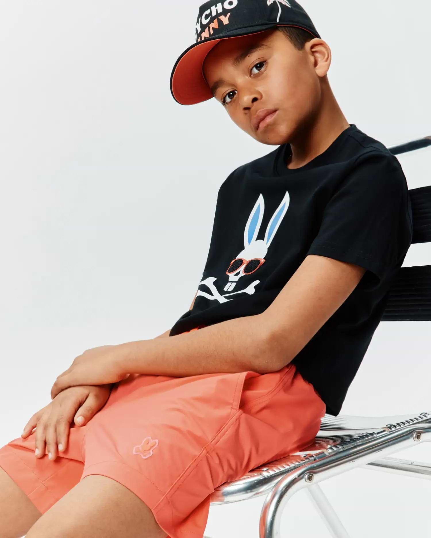 Black jersey t-shirt with large white Psycho Bunny skull logo, light blue ears, orange sunglasses. Paired with Kids Samos Hydrochromic Printed Swim Trunk in coral with small embroidered logo, and a black cap with orange brim.
