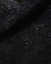 A detailed close-up of a Psycho Bunny KIDS PARKER HYDROCHROMIC SWIM TRUNK - B0W646C200 fabric displaying a subtle textured pattern with skull and crossbones and other abstract shapes. The slightly shiny, quick-dry material adds depth and dimension to the dark hydrochromatic pattern.