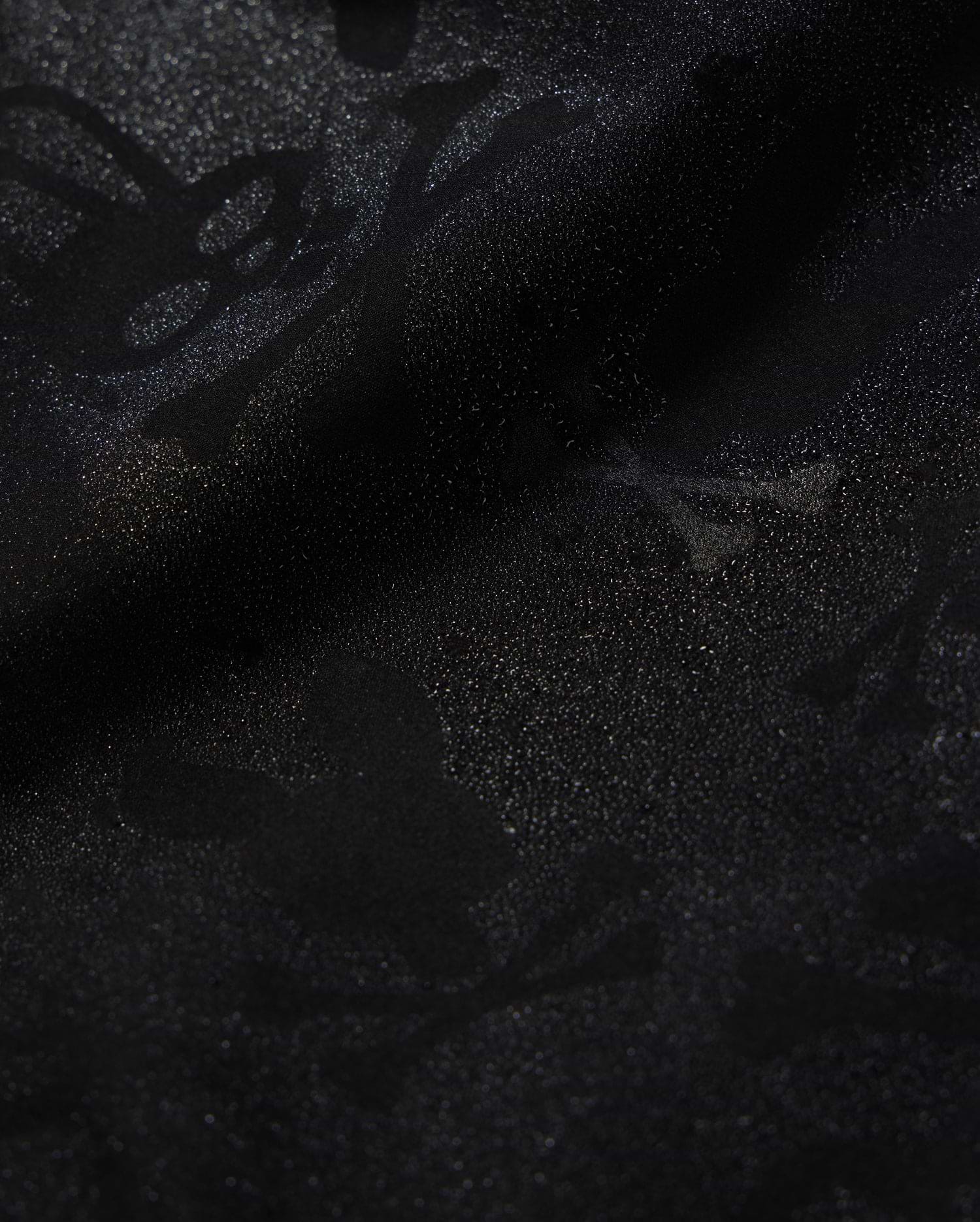 A detailed close-up of a Psycho Bunny KIDS PARKER HYDROCHROMIC SWIM TRUNK - B0W646C200 fabric displaying a subtle textured pattern with skull and crossbones and other abstract shapes. The slightly shiny, quick-dry material adds depth and dimension to the dark hydrochromatic pattern.