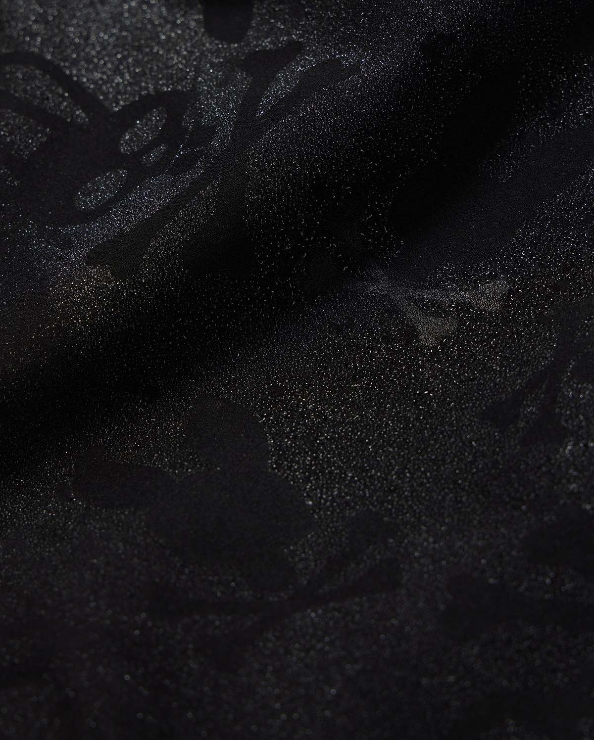 A detailed close-up of a Psycho Bunny KIDS PARKER HYDROCHROMIC SWIM TRUNK - B0W646C200 fabric displaying a subtle textured pattern with skull and crossbones and other abstract shapes. The slightly shiny, quick-dry material adds depth and dimension to the dark hydrochromatic pattern.