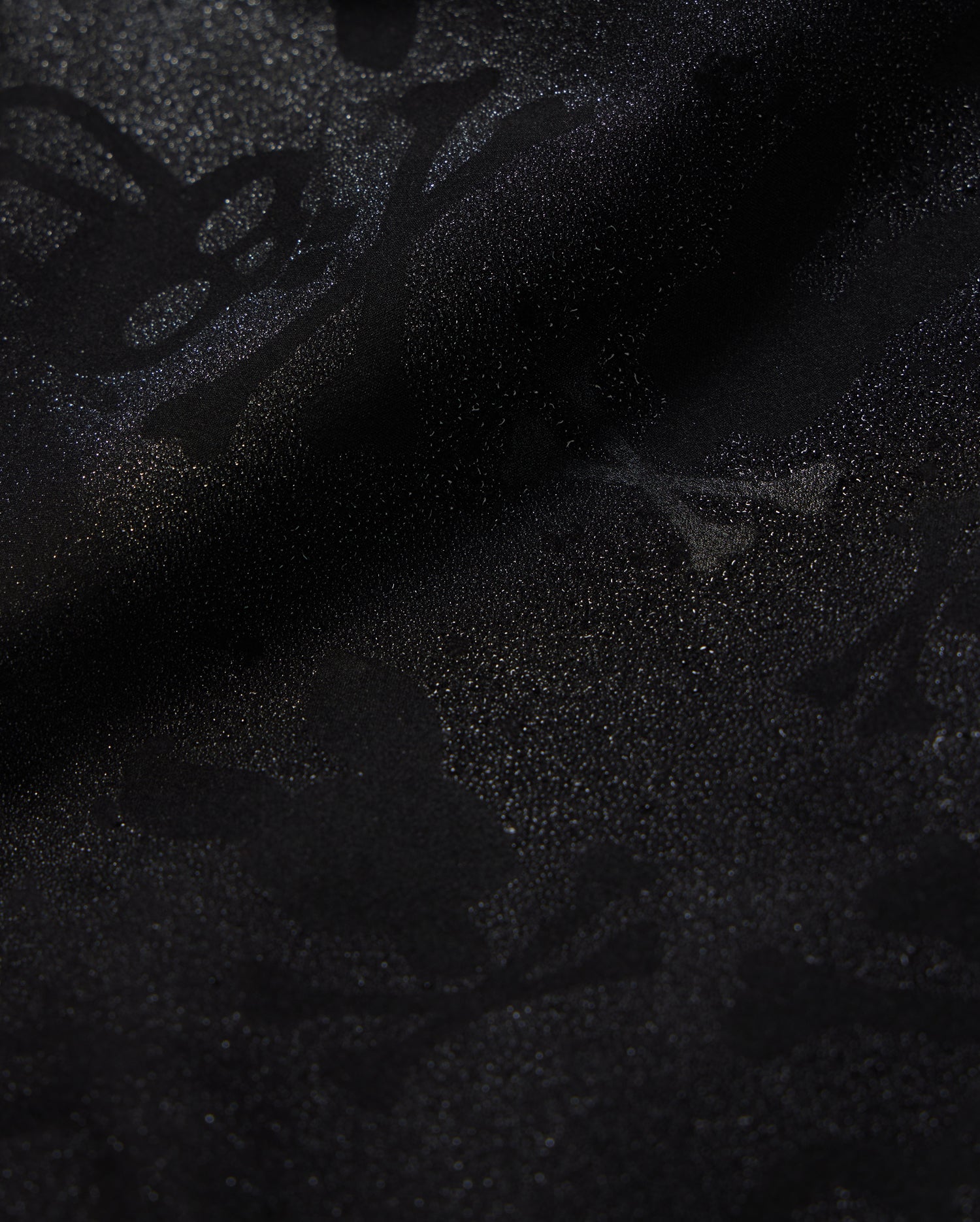 A detailed close-up of a Psycho Bunny KIDS PARKER HYDROCHROMIC SWIM TRUNK - B0W646C200 fabric displaying a subtle textured pattern with skull and crossbones and other abstract shapes. The slightly shiny, quick-dry material adds depth and dimension to the dark hydrochromatic pattern.