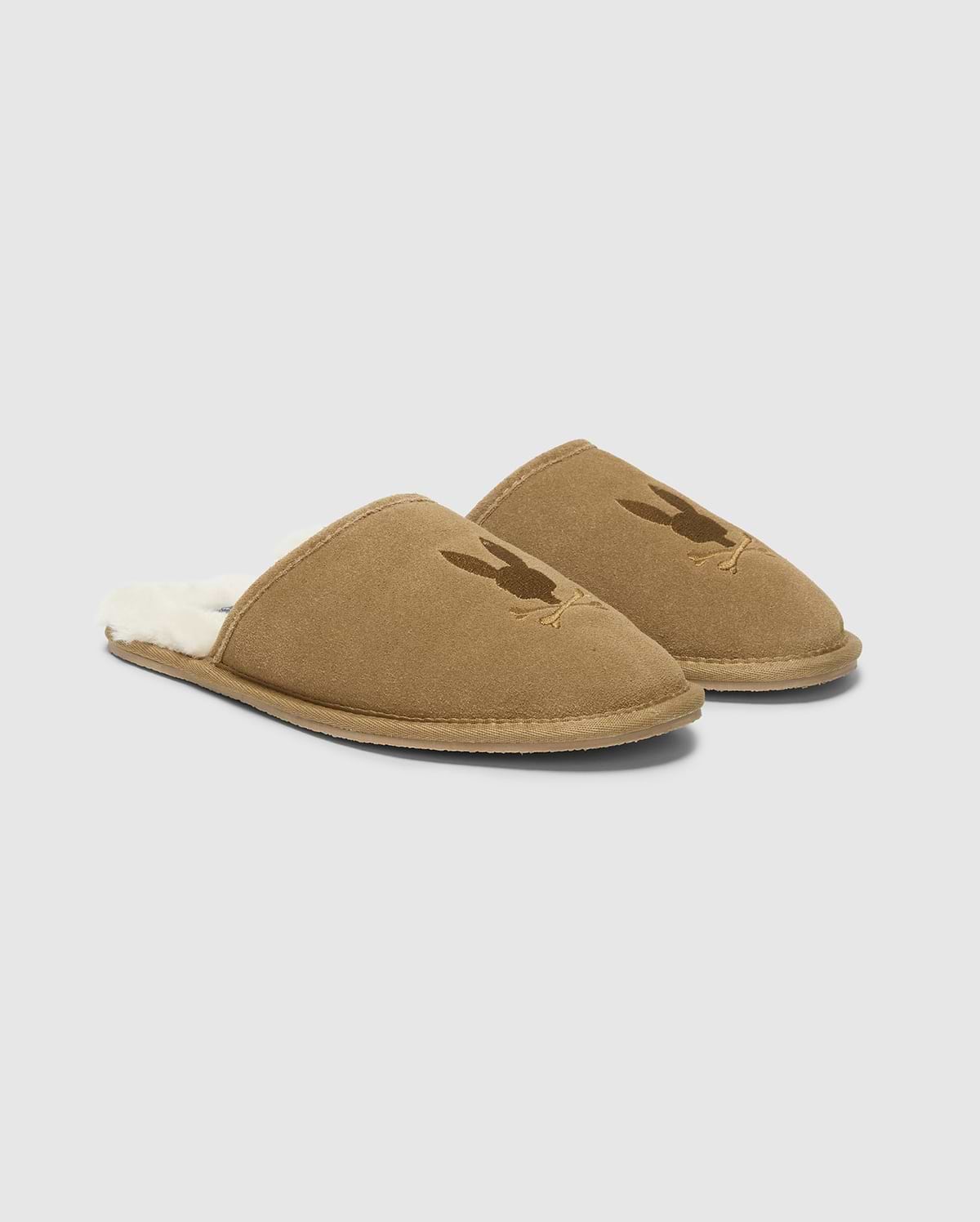 A pair of MENS SUEDE SLIPPERS by Psycho Bunny, crafted from cozy cow suede leather in brown with a soft, fuzzy interior. Each slipper is adorned with a subtle rabbit silhouette on the upper part, set against a clean white background. (1)