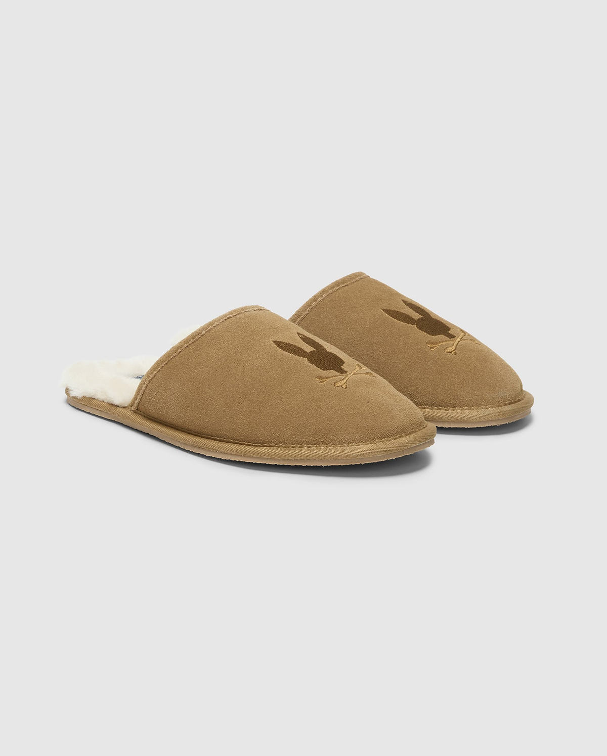 A pair of MENS SUEDE SLIPPERS by Psycho Bunny, crafted from cozy cow suede leather in brown with a soft, fuzzy interior. Each slipper is adorned with a subtle rabbit silhouette on the upper part, set against a clean white background. (1)