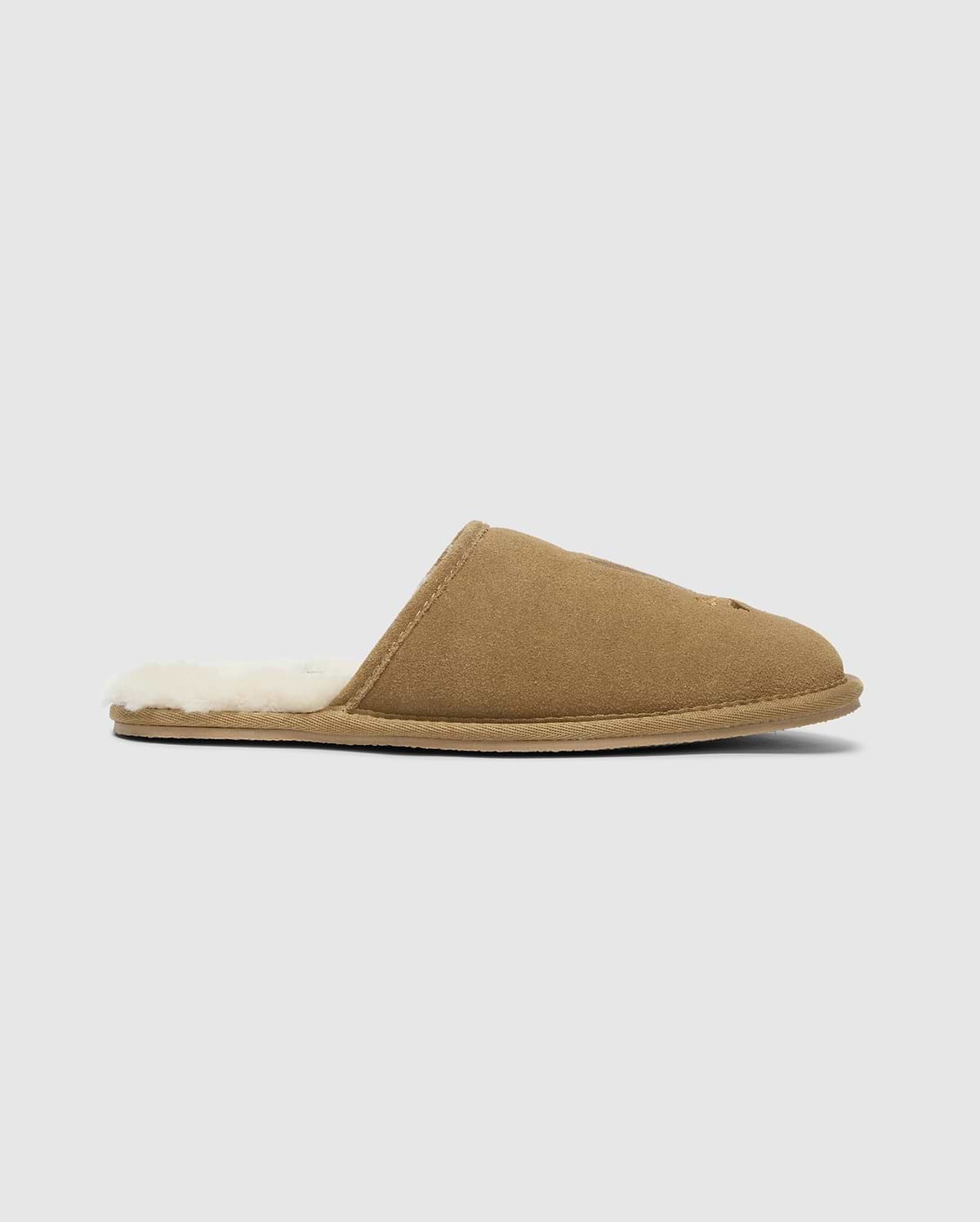 The MENS SUEDE SLIPPERS by Psycho Bunny, model B6A357E200, are crafted from brown cow suede leather and feature a plush off-white lining. These men's slippers have a flat sole and open back design, making them ideal for indoor comfort. The background is a subtle light gray. (2)