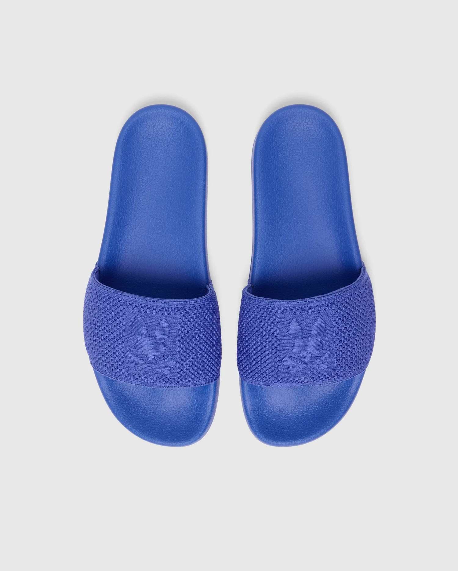 Royal blue MENS KNIT SLIDES with a textured footbed and a knitted strap featuring a large, tonal, signature Psycho Bunny skull logo woven into the fabric.