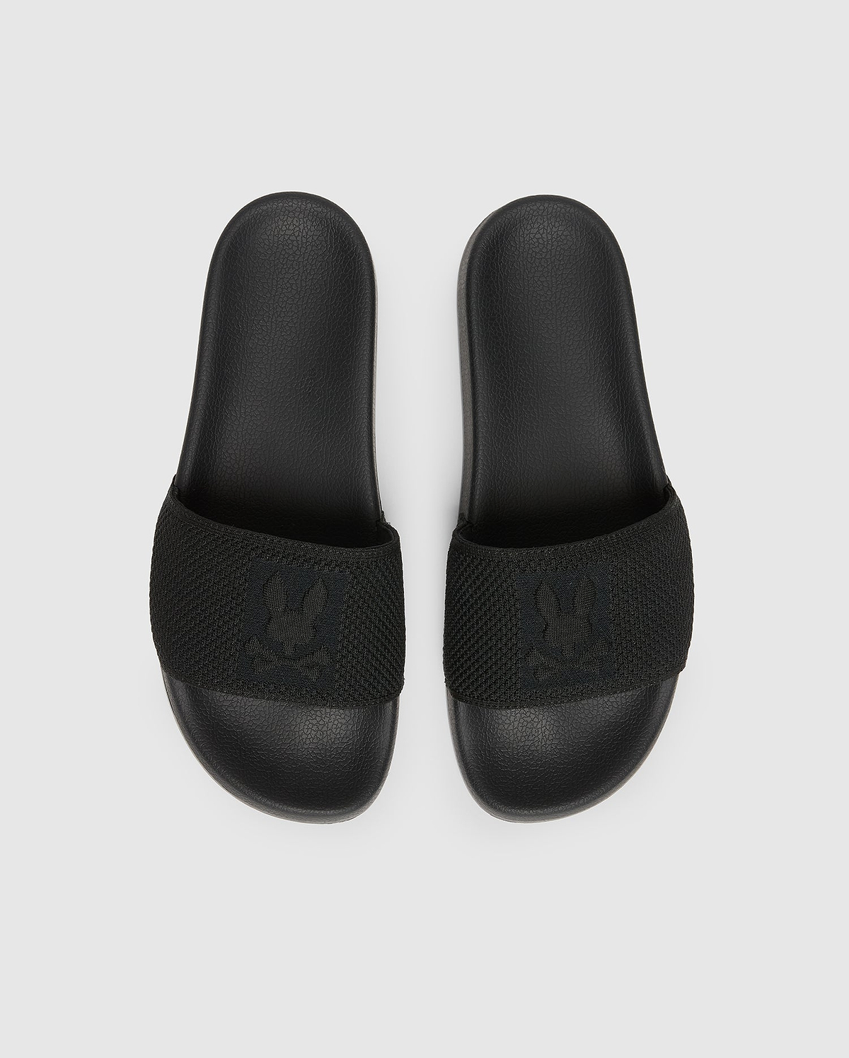 The Psycho Bunny MENS KNIT SLIDES - B6A474F200 are black slide sandals with textured straps and feature a flocked Bunny emblem design. They offer comfort with a molded footbed and are displayed on a plain light grey background.