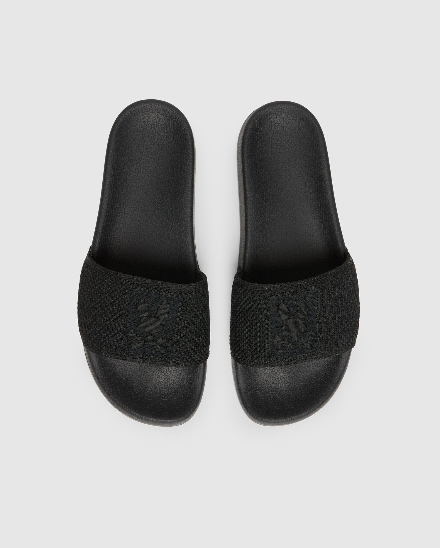 The Psycho Bunny MENS KNIT SLIDES - B6A474F200 are black slide sandals with textured straps and feature a flocked Bunny emblem design. They offer comfort with a molded footbed and are displayed on a plain light grey background.