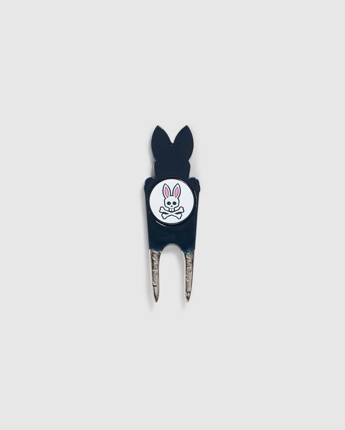 A Psycho Bunny DIVOT TOOL - B6A699C200 featuring a black handle with a white circular logo. The logo shows an illustration of a bunny head with crossed bones underneath. The two prongs of the tool are metal, and it includes a magnetic ball marker. Ideal for those seeking unique golf gifts.