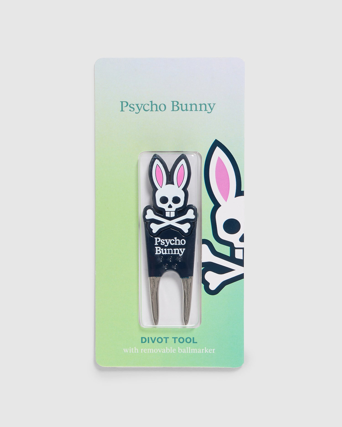 A Psycho Bunny branded DIVOT TOOL - B6A699C200 with a removable magnetic ball marker is displayed in its packaging. The tool features a bunny logo with crossbones and the text "Psycho Bunny" below it. The packaging has a green gradient background and a larger bunny logo on the right side, making it perfect for golf gifts.