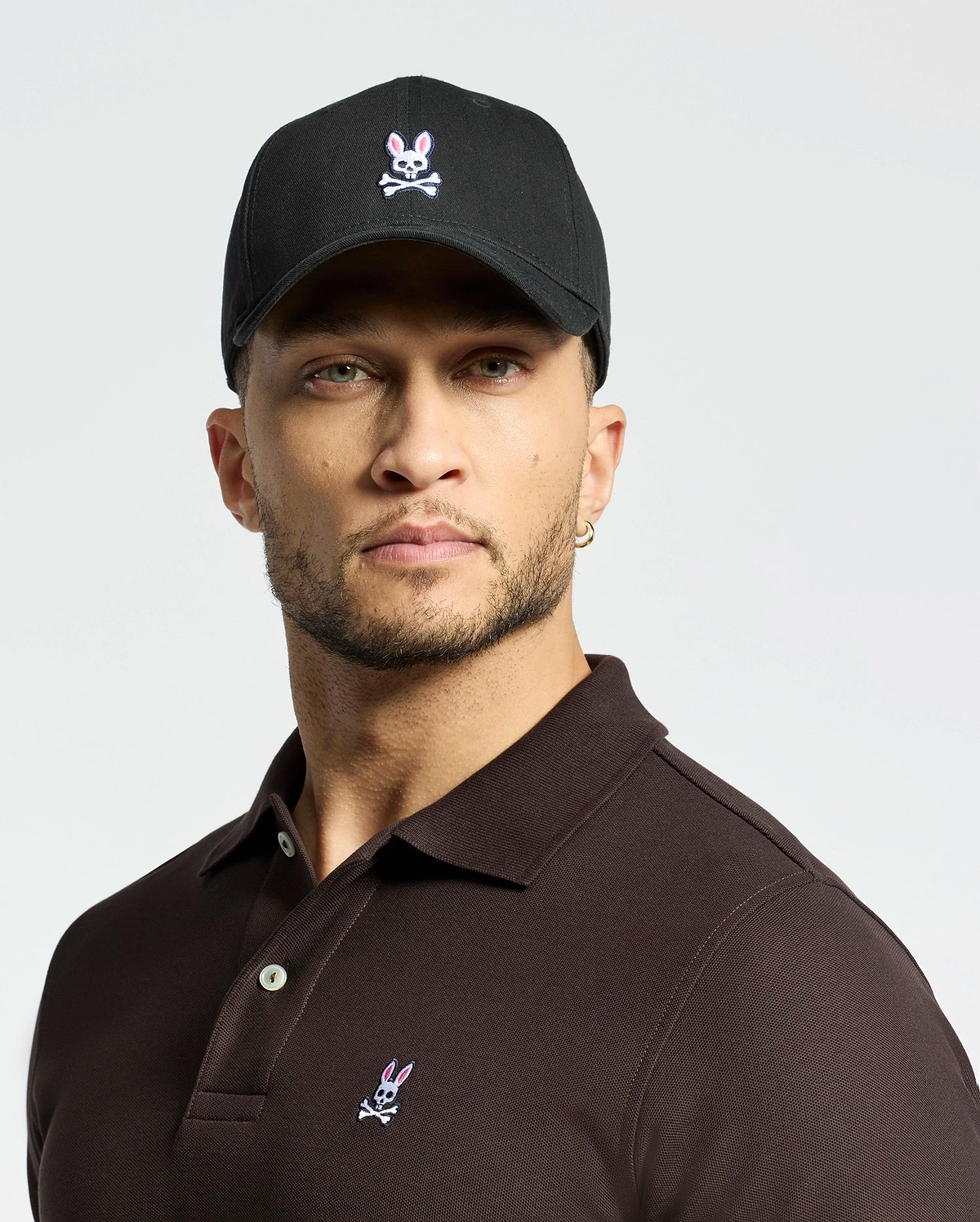 A person dons the MENS CLASSIC BASEBALL CAP by Psycho Bunny, featuring an embroidered Bunny logo. They pair it with a dark polo, possess a short beard and an earring, and pose against a light gray background, highlighting the adjustable fit of both items for comfort.