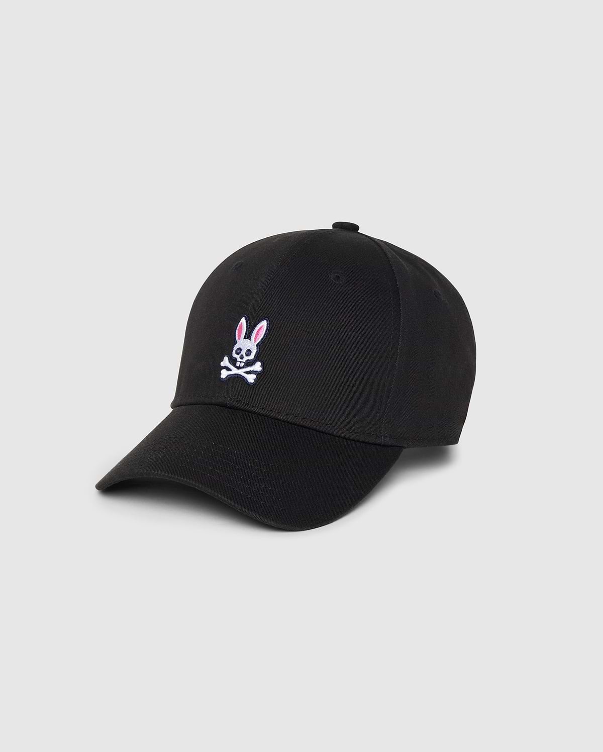 A Psycho Bunny MENS CLASSIC BASEBALL CAP - B6A816A1HT in black featuring a small embroidered pink and white bunny above crossed bones on the front. Made from durable cotton twill, it has a curved brim and six panels with eyelets for ventilation. The background is plain and light gray.