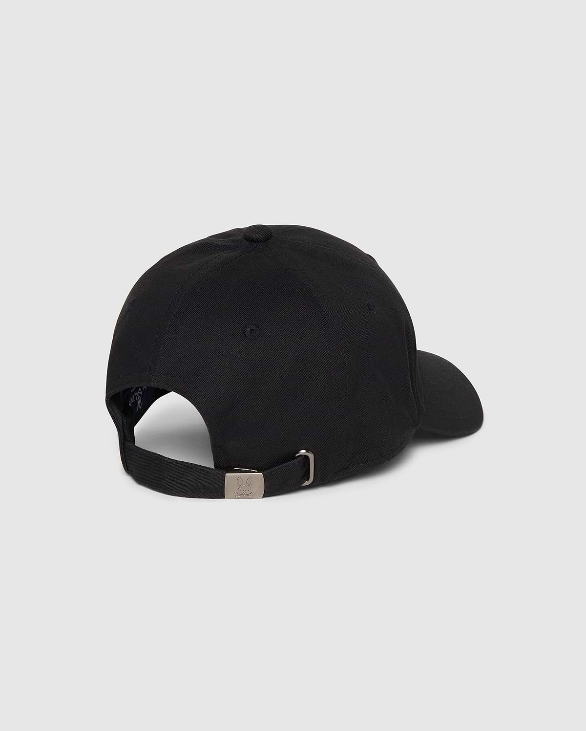 A Psycho Bunny MENS CLASSIC BASEBALL CAP - B6A816A1HT is shown from the back. The black cotton twill cap features an adjustable strap with a metal buckle and a small round vent hole on the side. The background is a plain grey.