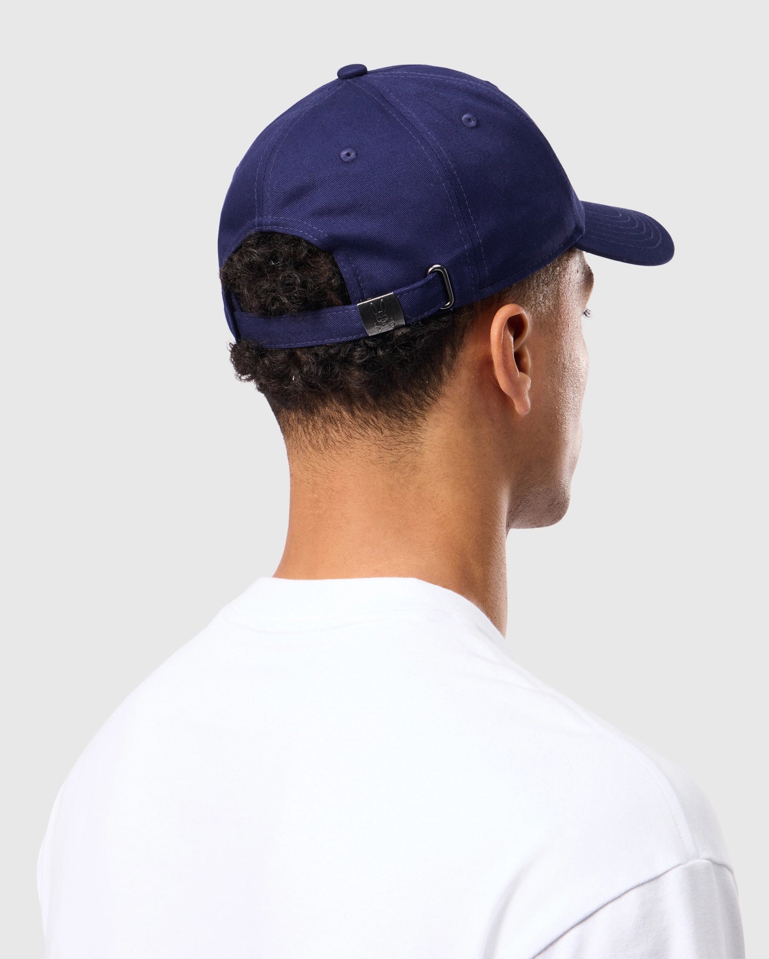 A person is seen from behind, wearing a timeless dark blue Psycho Bunny Men's Classic Baseball Cap - B6A816A1HT, adjusted with a buckle strap. They are also wearing a plain white shirt made from 100% cotton. The background is neutral gray.