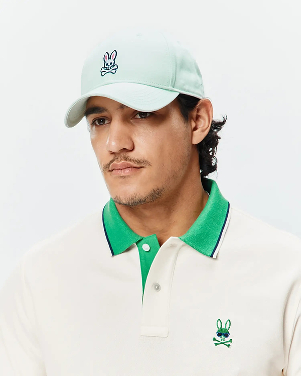 Off-white pique polo with bright green collar, navy stripe, and embroidered Psycho Bunny skull logo with sunglasses on chest. Light mint Mens Classic Baseball Cap with embroidered Psycho Bunny logo.
