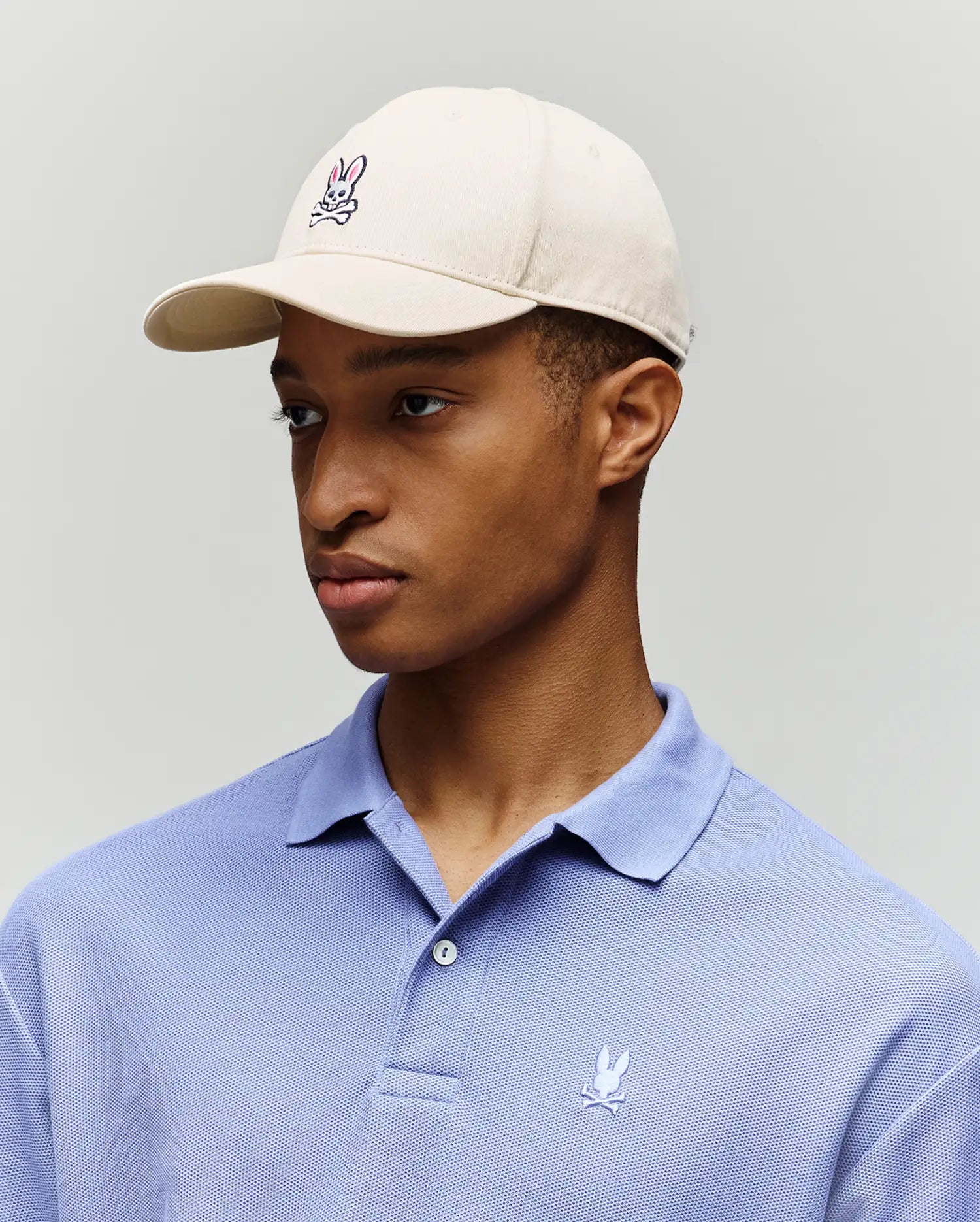 A person wearing a Psycho Bunny MENS CLASSIC BASEBALL CAP (B6A816A1HT) in beige and a light purple polo, both with the signature embroidered bunny and crossbones logo, stands against a plain light background.