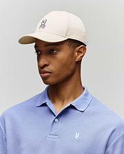 A person wearing a Psycho Bunny MENS CLASSIC BASEBALL CAP (B6A816A1HT) in beige and a light purple polo, both with the signature embroidered bunny and crossbones logo, stands against a plain light background.