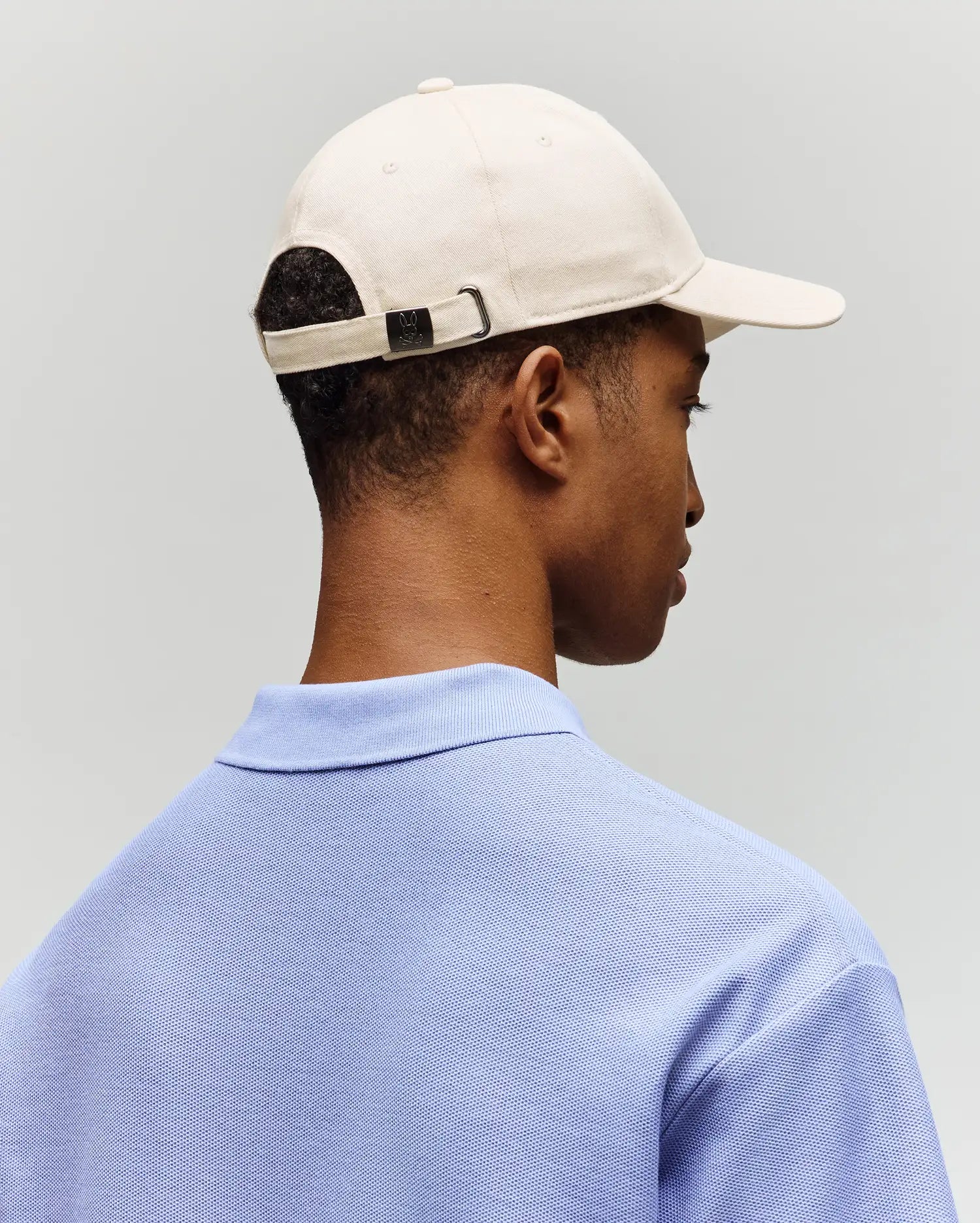 A person is shown from behind wearing the Psycho Bunny MENS CLASSIC BASEBALL CAP - B6A816A1HT in light beige and a light blue collared shirt, set against a plain light gray background.