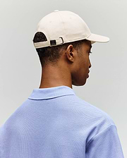 A person is shown from behind wearing the Psycho Bunny MENS CLASSIC BASEBALL CAP - B6A816A1HT in light beige and a light blue collared shirt, set against a plain light gray background.