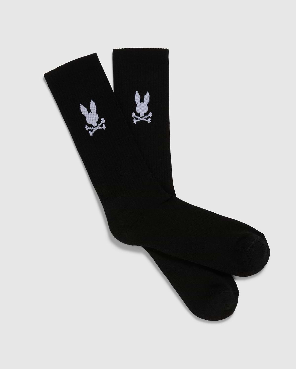 A pack of 3 pairs of black men's knee-high sport socks from the Psycho Bunny brand, featuring a white skull and crossbones symbol with bunny ears on each sock. Made from luxurious Peruvian pima cotton, these MENS 3 PACK SPORT SOCK - B6F997D200 socks are displayed positioned overlapping each other against a plain, light gray background.