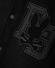 Close-up of the Psycho Bunny MENS BRAYDEN WOOL LEATHER VARSITY BOMBER - B6J876D200, a black wool-blend bomber jacket adorned with large, textured "D" and "B" letters, along with a stitched emblem showcasing a stylized rabbit head and crossbones in dark fabric. The image emphasizes the intricate detailing and craftsmanship characteristic of a classic varsity jacket.