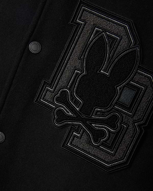 Close-up of the Psycho Bunny MENS BRAYDEN WOOL LEATHER VARSITY BOMBER - B6J876D200, a black wool-blend bomber jacket adorned with large, textured "D" and "B" letters, along with a stitched emblem showcasing a stylized rabbit head and crossbones in dark fabric. The image emphasizes the intricate detailing and craftsmanship characteristic of a classic varsity jacket.