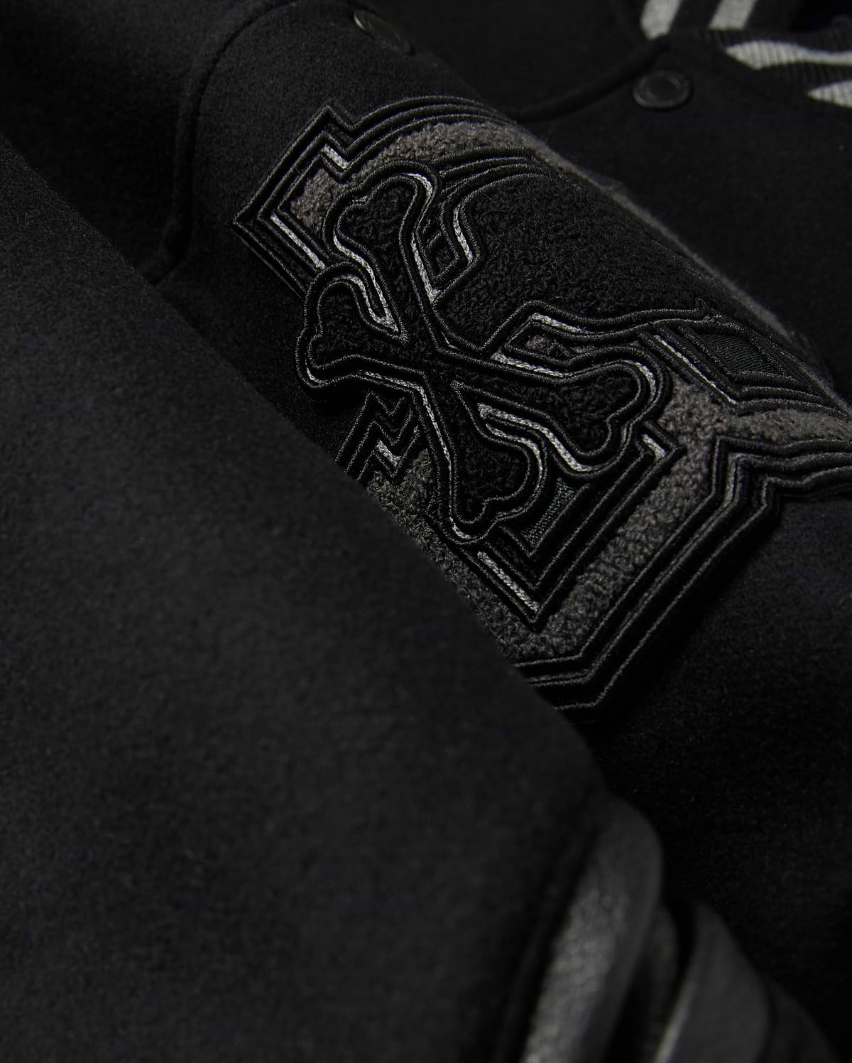 Close-up of the Psycho Bunny MENS BRAYDEN WOOL LEATHER VARSITY BOMBER - B6J876D200, showcasing an embroidered design with overlapping bones and intricate stitching. The thick, textured wool-blend fabric highlights the detailed craftsmanship of this black varsity jacket.