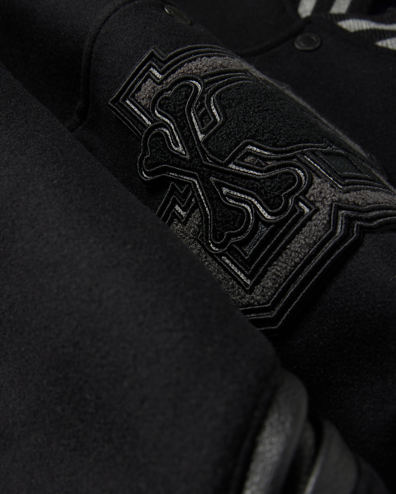 Close-up of the Psycho Bunny MENS BRAYDEN WOOL LEATHER VARSITY BOMBER - B6J876D200, showcasing an embroidered design with overlapping bones and intricate stitching. The thick, textured wool-blend fabric highlights the detailed craftsmanship of this black varsity jacket.