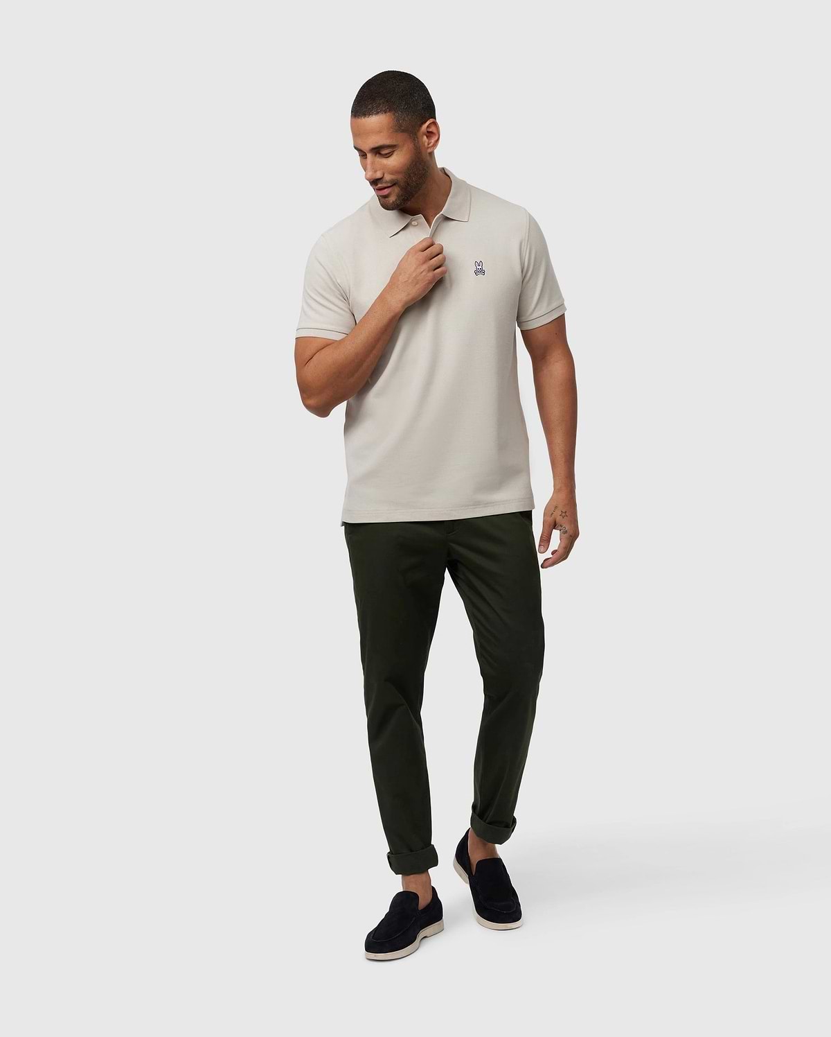 A man wearing a timeless men's Psycho Bunny MENS CLASSIC PIQUE POLO SHIRT - B6K001B200 in light grey, dark green pants, and black loafers stands against a plain white background. He looks down and to his right while adjusting the collar of his luxurious Pima cotton polo with his right hand.