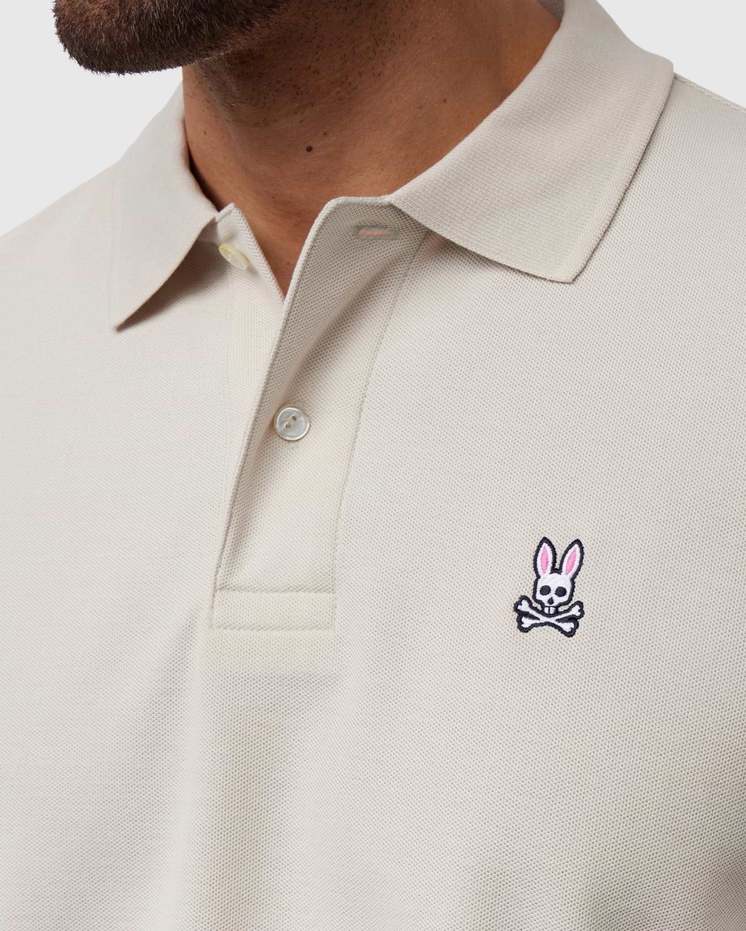 Close-up shot of a person wearing a light beige Psycho Bunny MENS CLASSIC PIQUE POLO SHIRT - B6K001B200 with a collar and buttons. This timeless shirt, crafted from luxurious Pima cotton, features an embroidered logo on the left chest area depicting a pink bunny head above crossed bones. Only part of the person’s face and neck are visible.