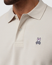 Close-up shot of a person wearing a light beige Psycho Bunny MENS CLASSIC PIQUE POLO SHIRT - B6K001B200 with a collar and buttons. This timeless shirt, crafted from luxurious Pima cotton, features an embroidered logo on the left chest area depicting a pink bunny head above crossed bones. Only part of the person’s face and neck are visible.