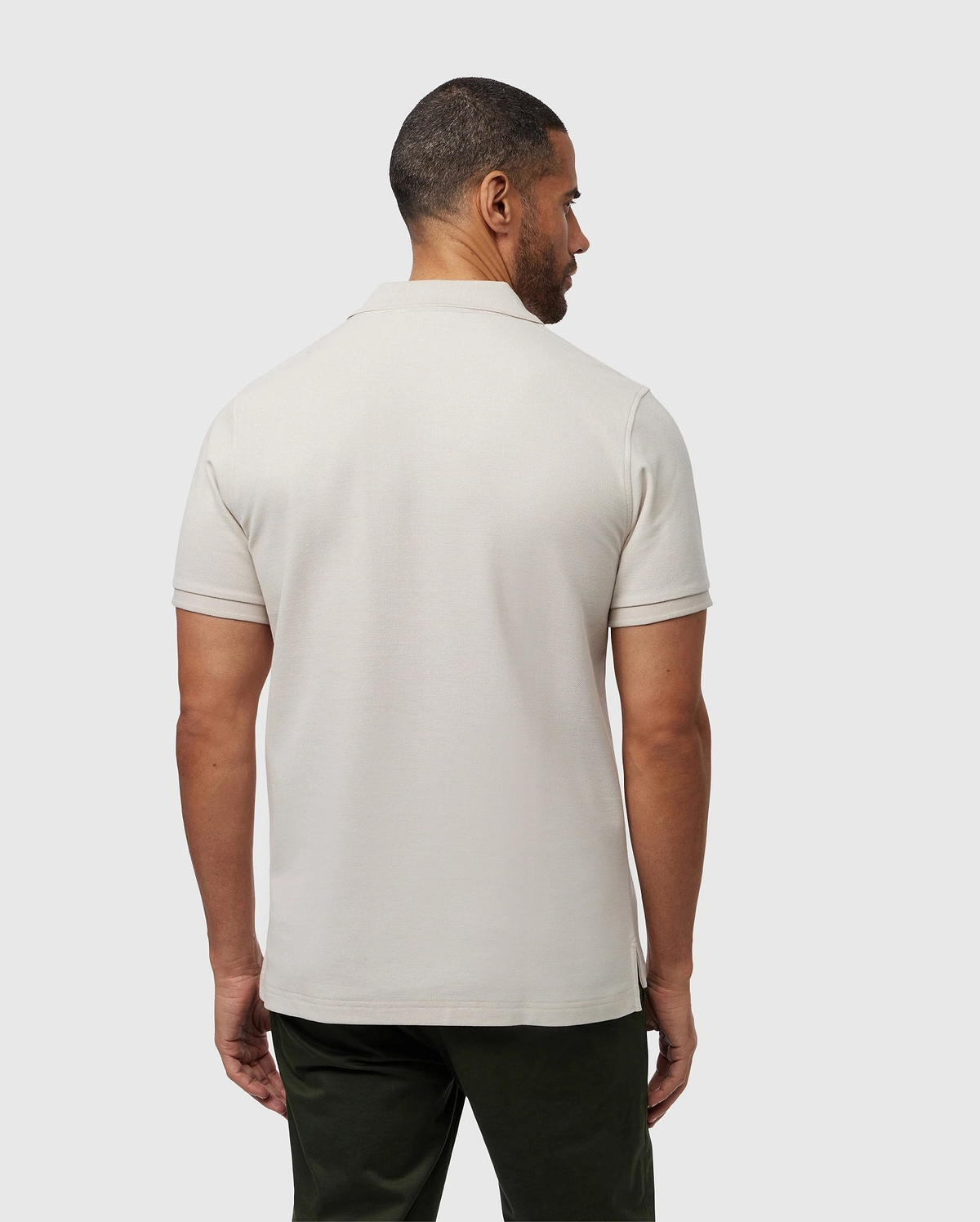 A man with short hair is standing with his back to the camera, showcasing the perfect fit of his light beige MENS CLASSIC PIQUE POLO SHIRT - B6K001B200 by Psycho Bunny. He pairs it effortlessly with dark green pants against a plain, light gray background.