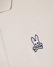 Close-up of a beige textured fabric from the Psycho Bunny MENS CLASSIC PIQUE POLO SHIRT - B6K001B200, with an embroidered logo featuring a white skull and crossbones with pink-eared bunny ears on the chest area. Crafted from luxurious Pima cotton for a perfect fit.
