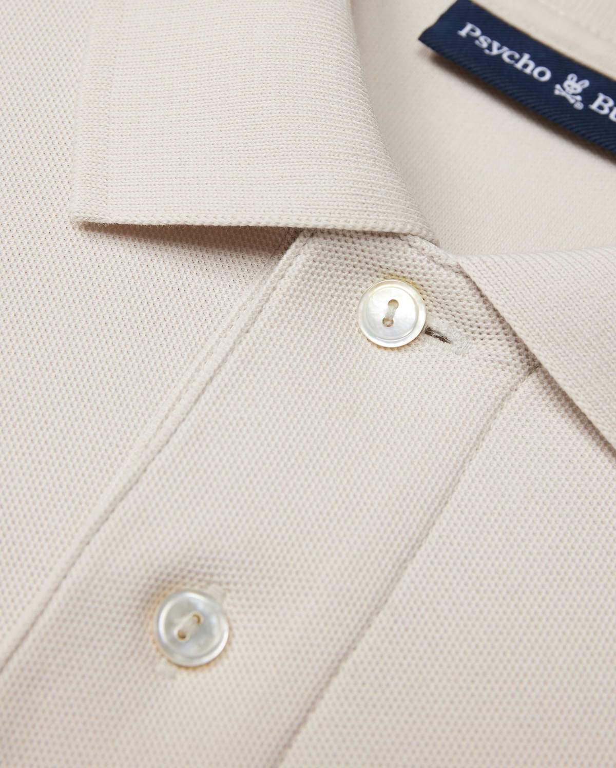 Close-up of a beige Psycho Bunny MENS CLASSIC PIQUE POLO SHIRT - B6K001B200 collar with two white buttons. The timeless shirt fabric has a textured, waffle-like pattern. A navy blue label with white text is partially visible near the collar, hinting at its luxurious Pima cotton composition.
