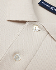 Close-up of a beige Psycho Bunny MENS CLASSIC PIQUE POLO SHIRT - B6K001B200 collar with two white buttons. The timeless shirt fabric has a textured, waffle-like pattern. A navy blue label with white text is partially visible near the collar, hinting at its luxurious Pima cotton composition.
