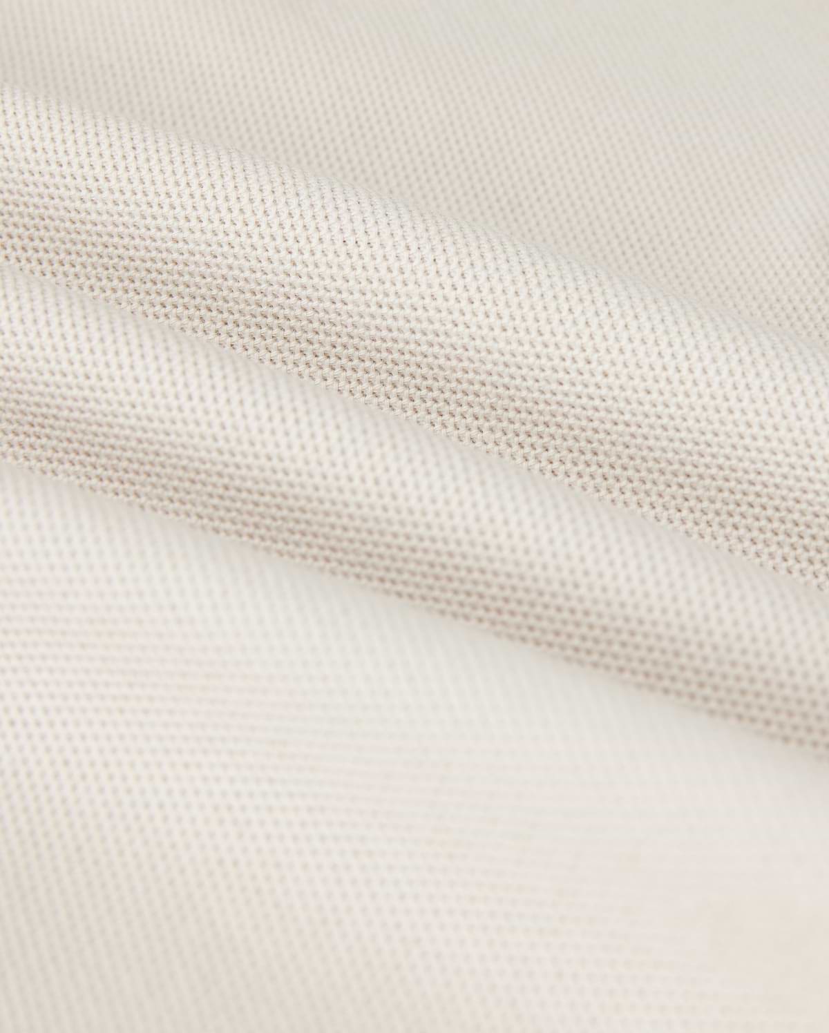 Close-up of a softly folded, light beige textured fabric with a subtle grid pattern, showcasing its smooth and pliable nature. The gentle folds add depth and highlight the luxurious Pima cotton's fine weave and soft sheen, hinting at the perfect fit for a Psycho Bunny Men's Classic Pique Polo Shirt - B6K001B200.