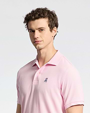 A person with curly hair wears a pale pink Psycho Bunny MENS CLASSIC PIQUE POLO SHIRT - B6K001CRPC, featuring a small embroidered logo on the chest, against a neutral background. The Pima cotton adds comfort as they look toward the camera with a slight smile.