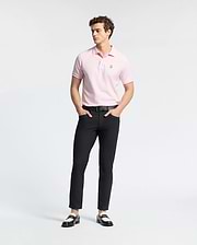 A person stands wearing a light pink Psycho Bunny MENS CLASSIC PIQUE POLO SHIRT - B6K001CRPC and black pants, hands in pockets. Two-tone loafers complete the look against a white background, highlighting the Pima cotton clothing.
