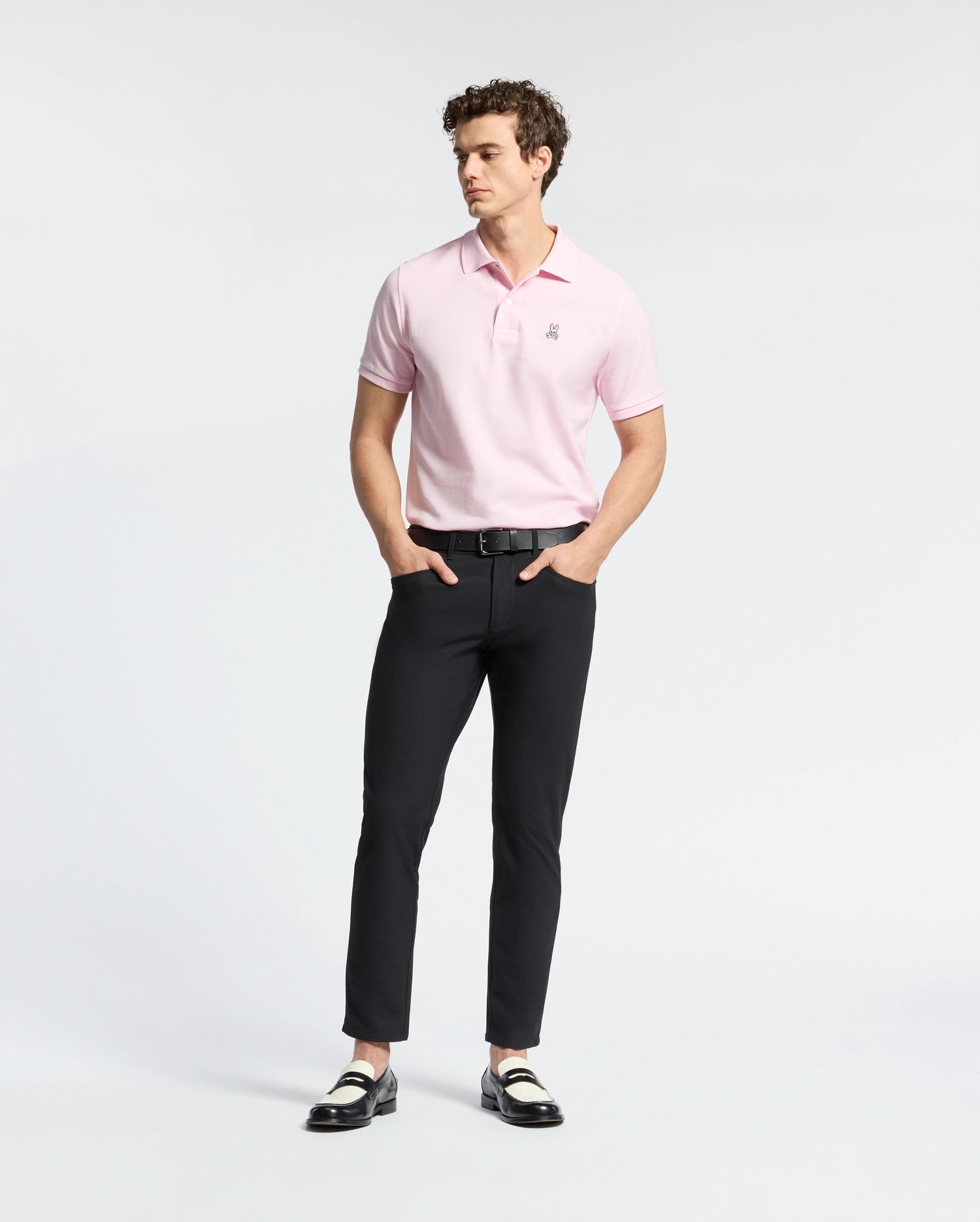 A person stands wearing a light pink Psycho Bunny MENS CLASSIC PIQUE POLO SHIRT - B6K001CRPC and black pants, hands in pockets. Two-tone loafers complete the look against a white background, highlighting the Pima cotton clothing.